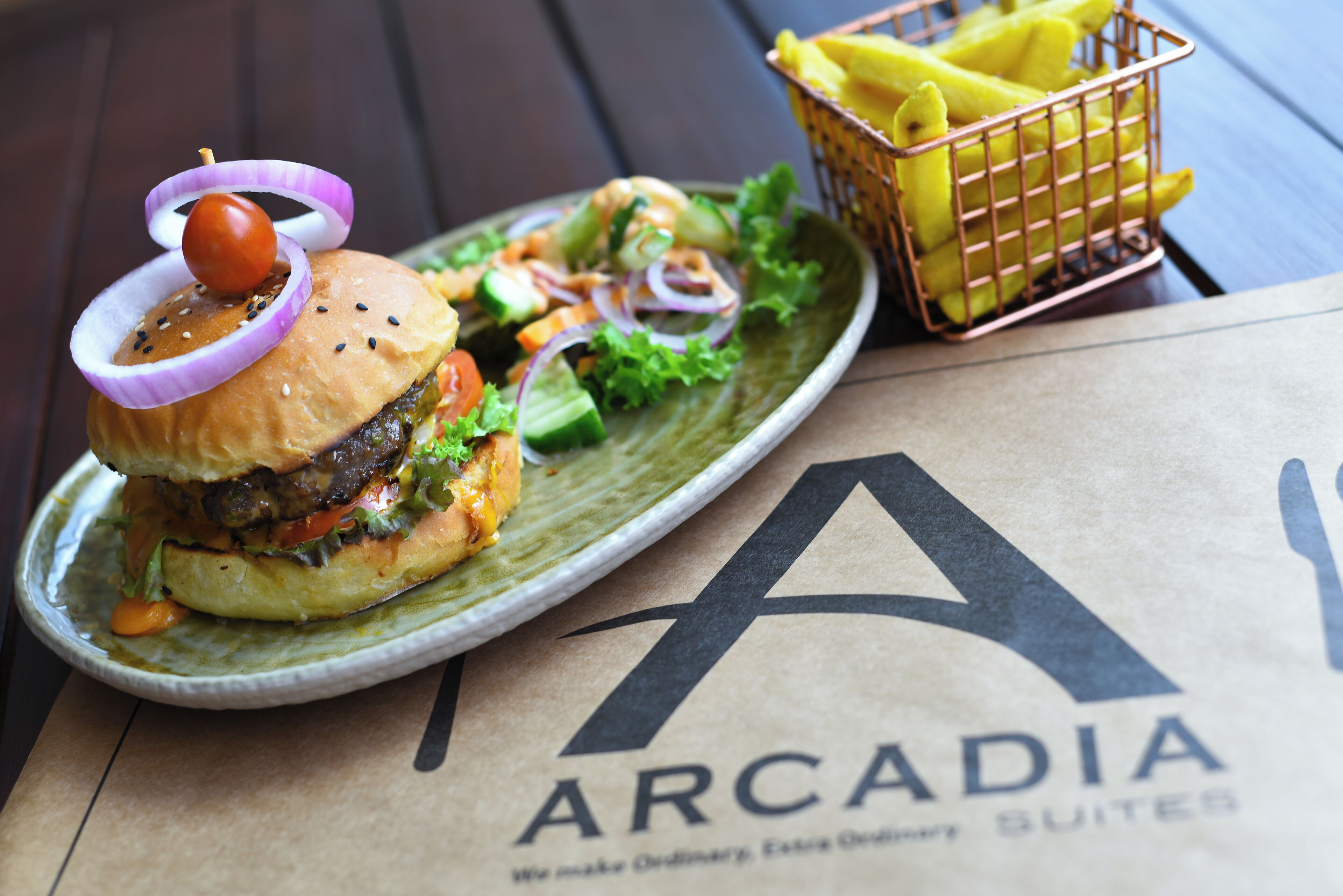 Ein Burger mit einer Kirsche und Zwiebelringen auf einem Teller, dazu ein Salat und Pommes, auf einem Tisch mit einer Tasche mit dem Aufdruck 'Arcadia'.