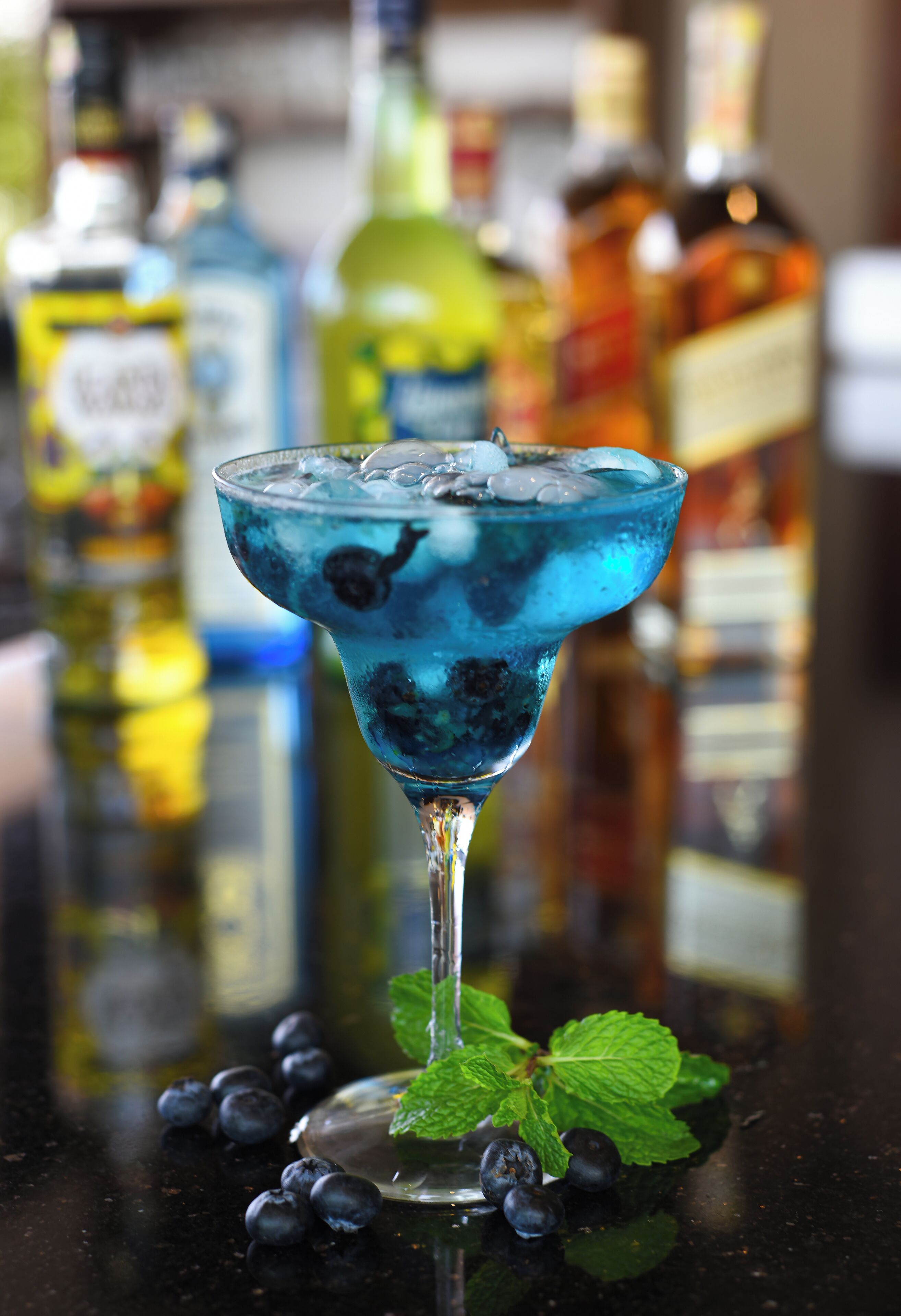A cocktail in a glass with ice cubes and a mint leaf garnish, with blurred bottles in the background.