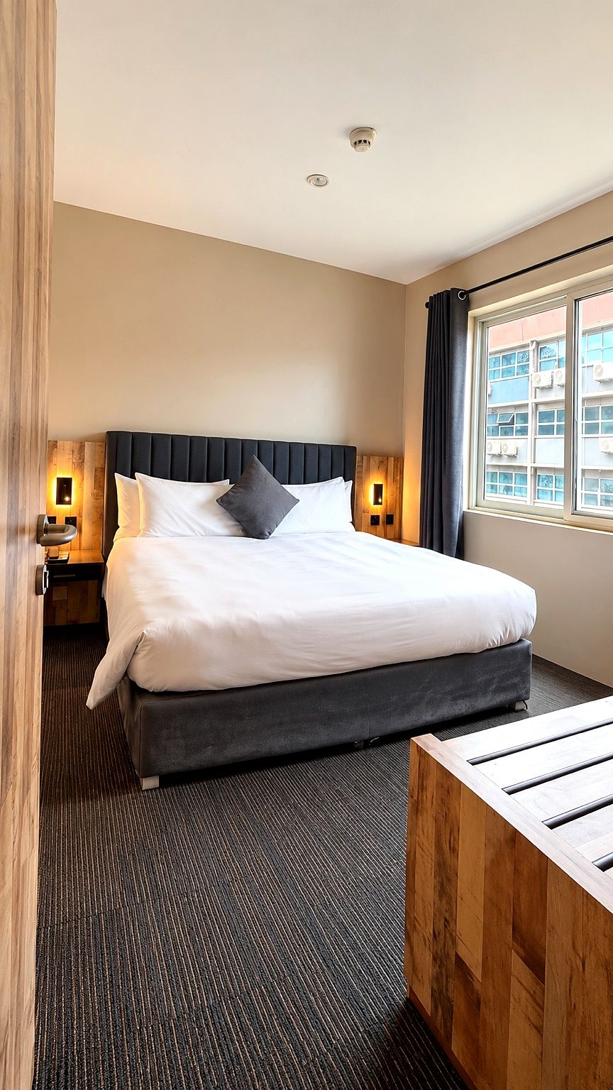 A hotel room with a large bed, two bedside lamps, a window with curtains, and a wooden bench.