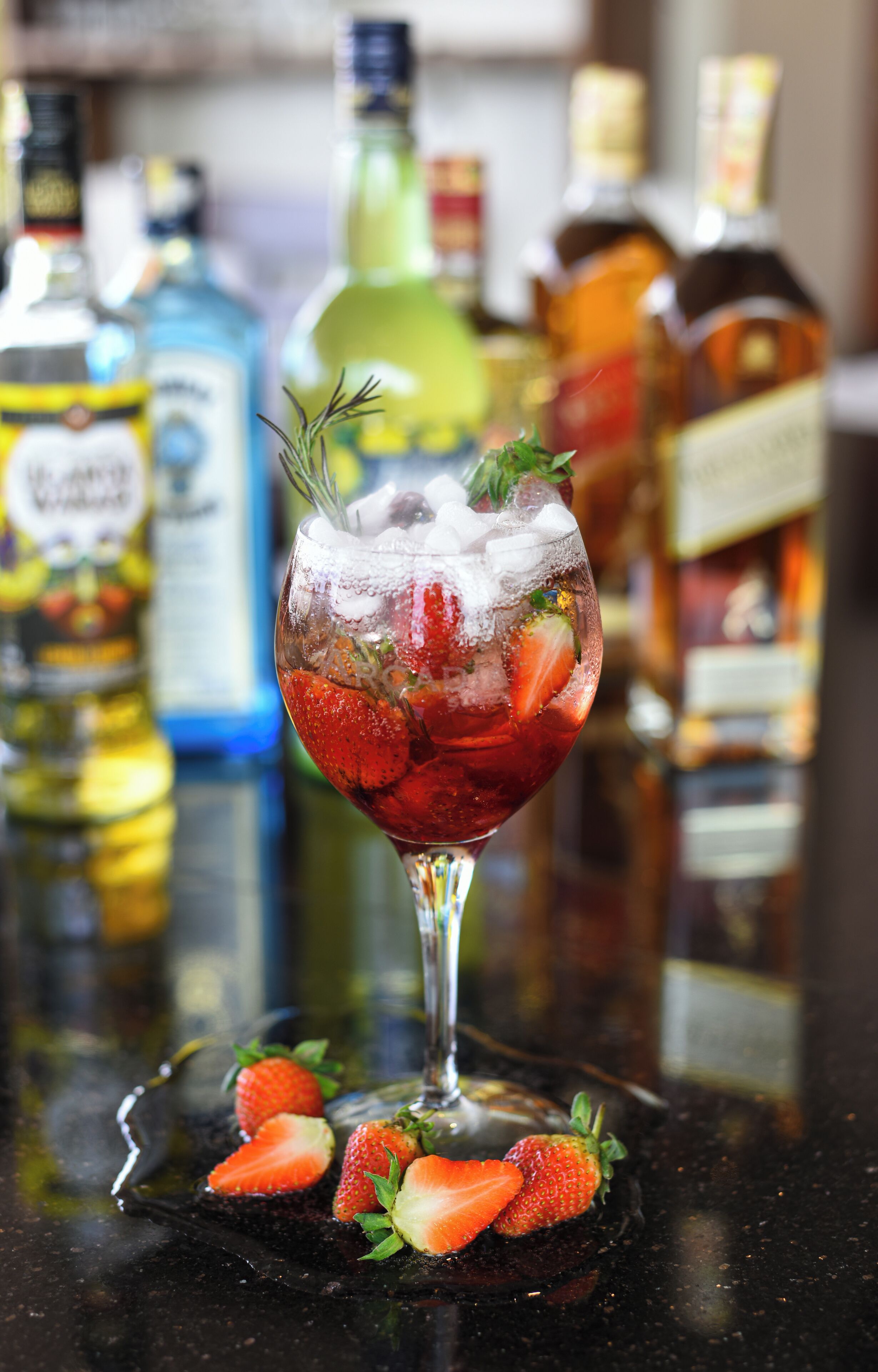 Um copo de cocktail com morangos e um ramo de alecrim sobre um apoio de mesa preto.