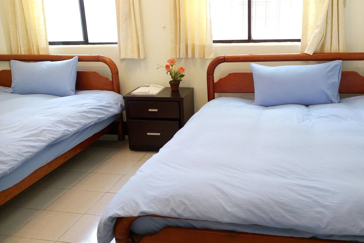 A hotel room with two single beds, a small bedside table with a lamp, and a window with curtains.
