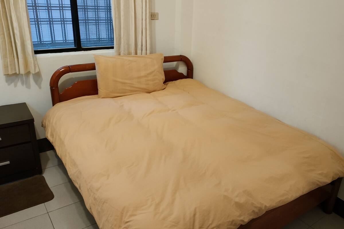 A single bed with a wooden headboard and a beige bedspread in a room with a window, a small bedside table, and a power outlet on the wall.
