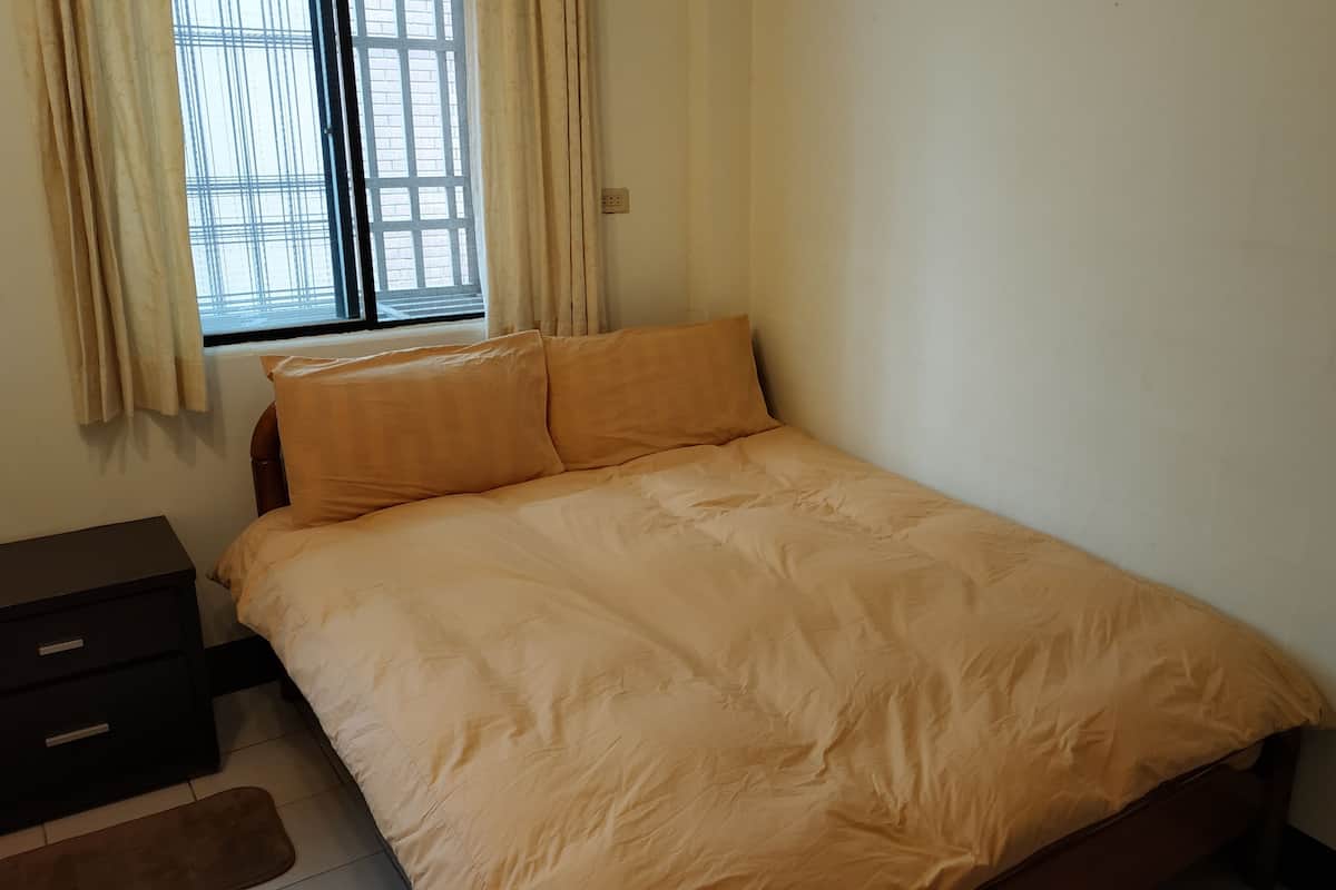 A bedroom with a bed, a nightstand, a window with curtains, and a wall socket.