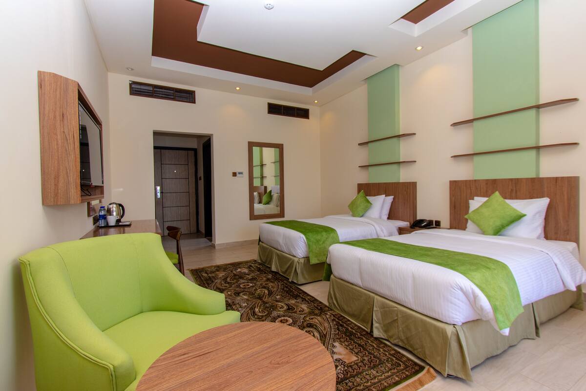 A hotel room with two beds, a green armchair, a wooden coffee table, and a patterned rug.