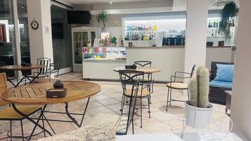 Un café con mesas redondas de madera y sillas de metal negro, un expositor de vidrio con aperitivos, y una zona de bar con varias botellas.