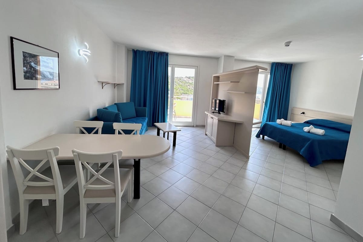 A modern hotel room with a dining area, a sofa, a TV, and two beds.