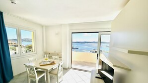 A room with a dining table, chairs, a TV, and a balcony with a view of the sea.