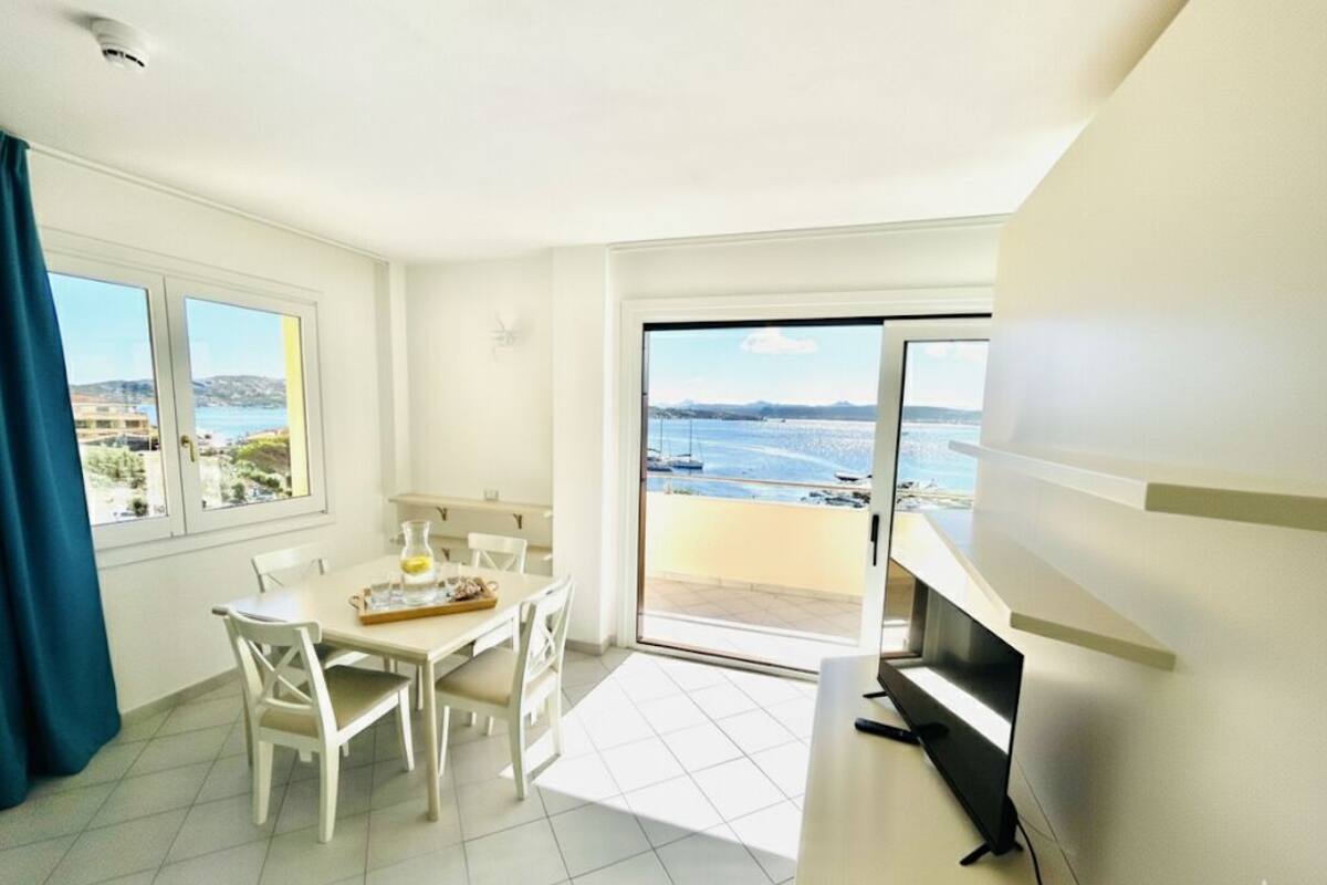 A room with a dining table, chairs, a TV, and a balcony with a view of the sea.