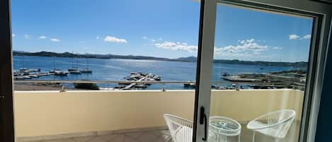 Un balcón con vistas despejadas a un puerto deportivo y embarcaciones, dos sillas blancas y un ventanal de vidrio.