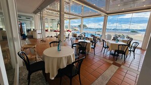 A restaurant with round tables and chairs, large windows offering a view of the sea, and a tiled floor.