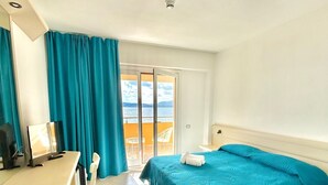 A hotel room with a bed, a desk, a chair, a computer, and a view of the sea through a sliding door.