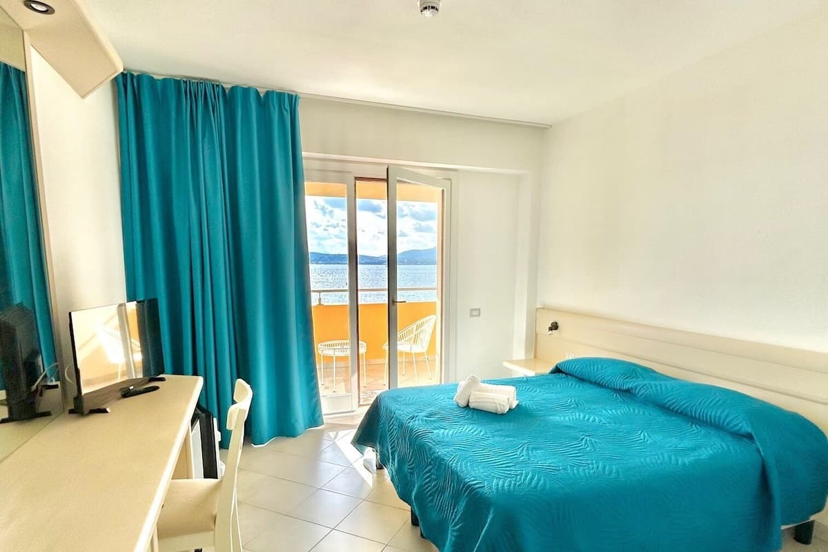 A hotel room with a bed, a desk, a chair, a computer, and a view of the sea through a sliding door.