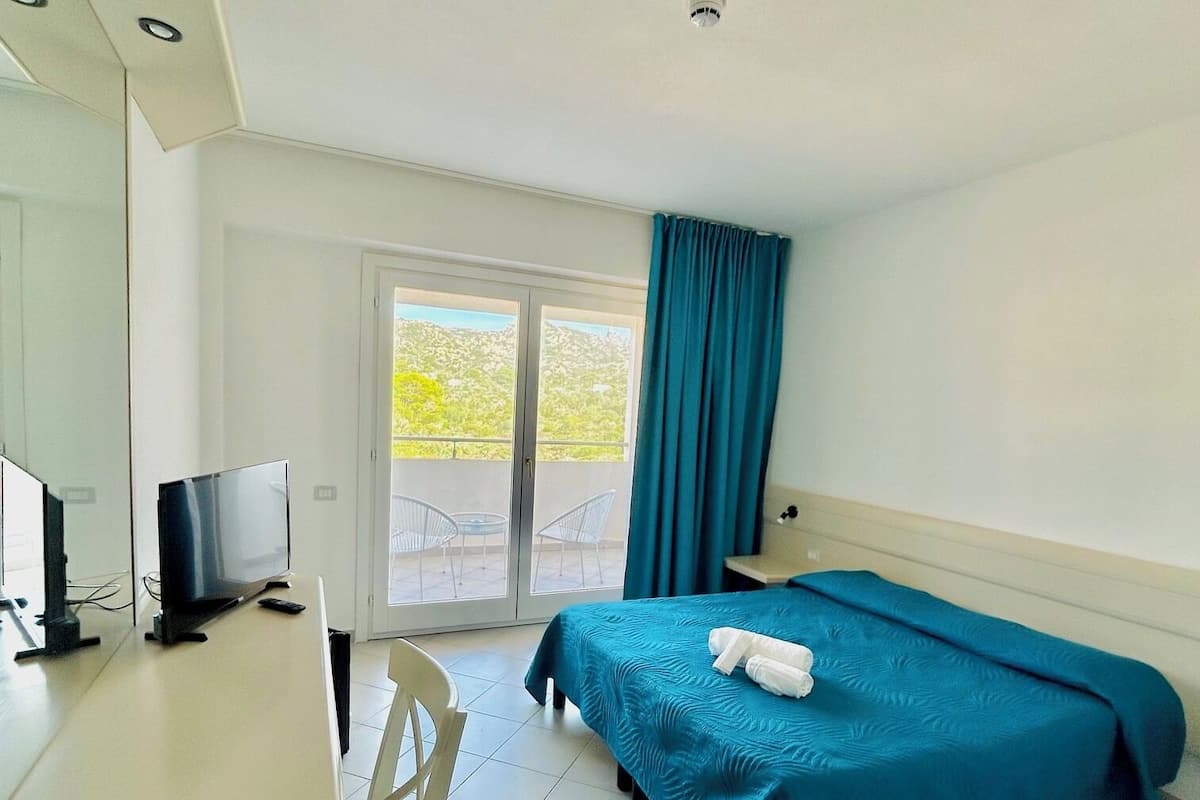 A hotel room with a bed, a desk with a computer, a chair, a TV, and a window with curtains.