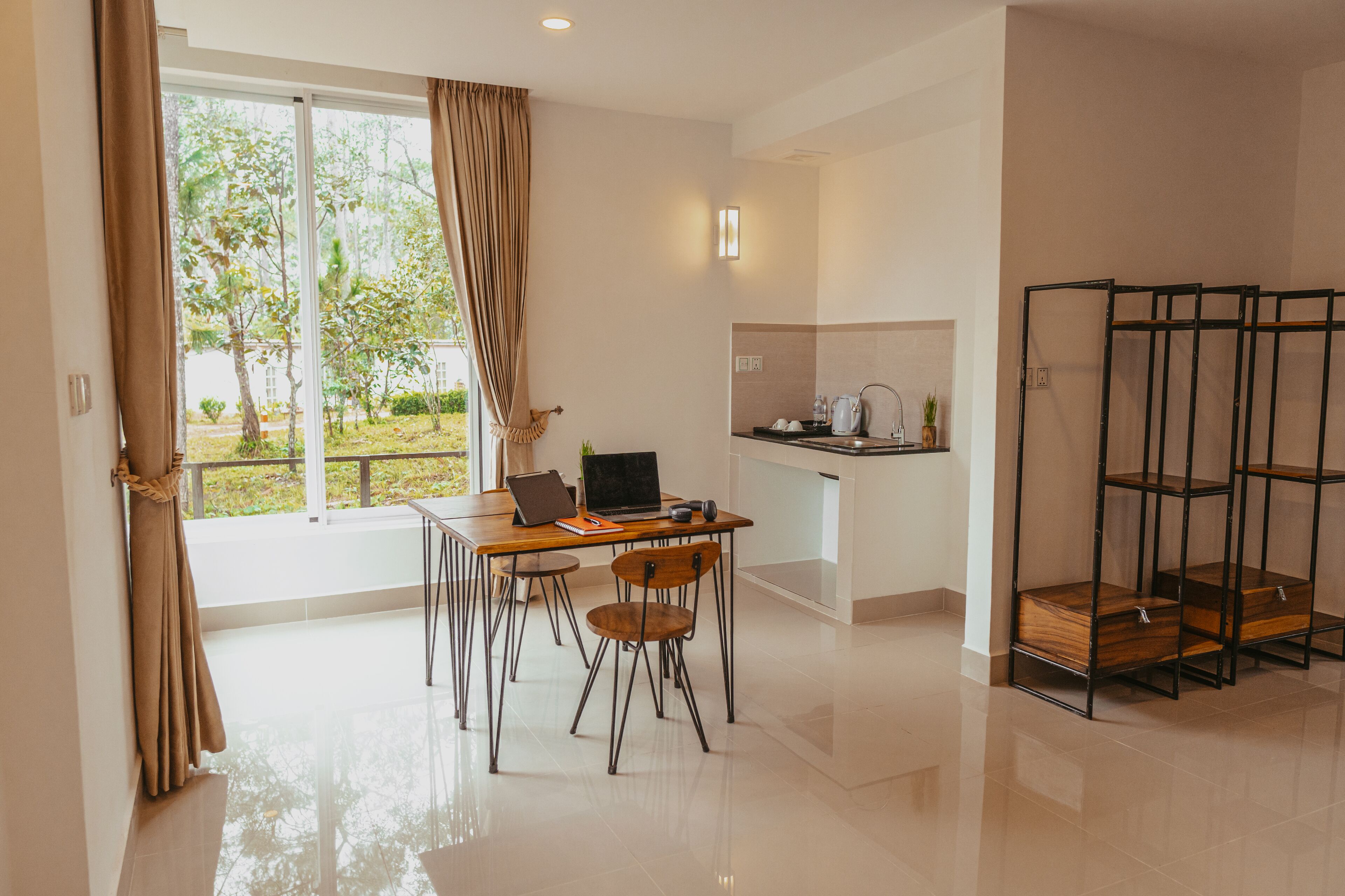 A modern kitchen with a dining area, a large window with curtains, and a view of greenery outside.