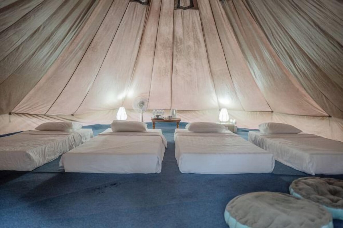 Una carpa acogedora con cuatro camas dispuestas en fila, una mesita y dos lámparas.