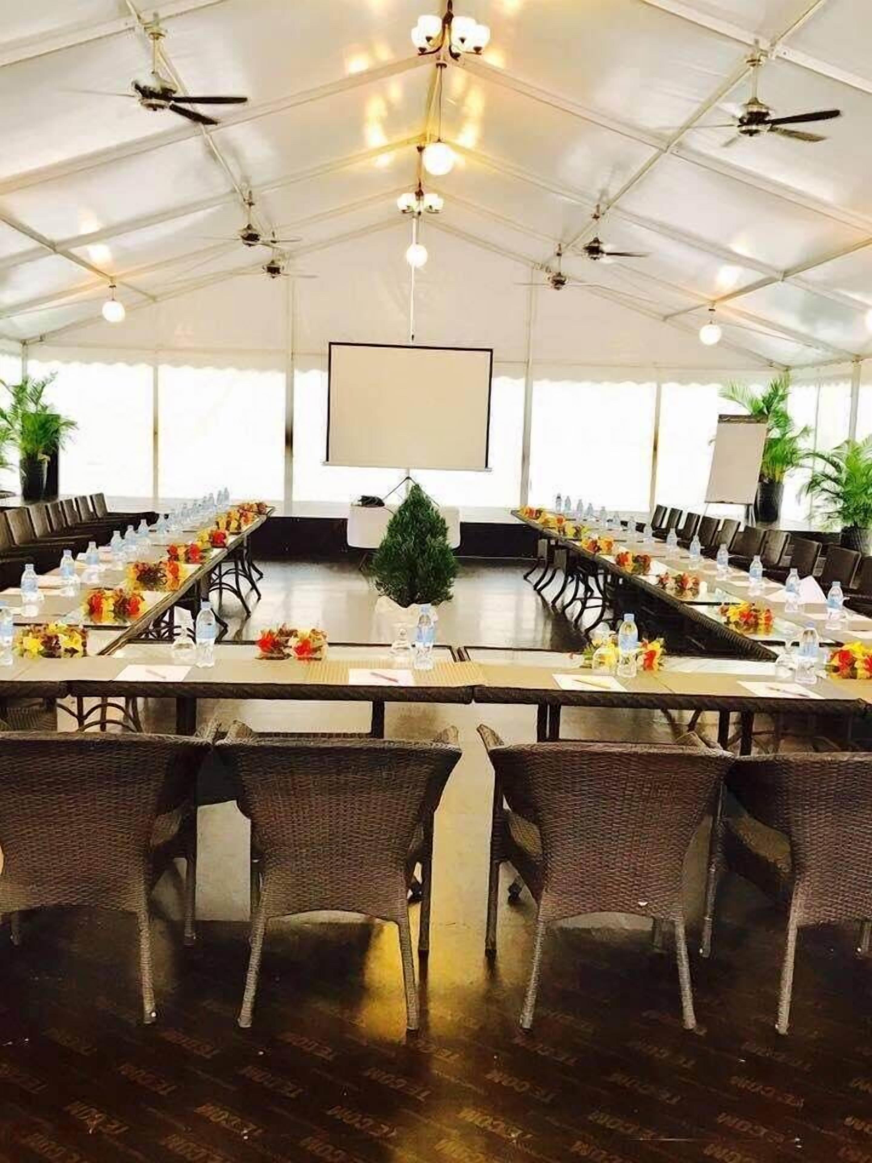 A conference room with a long table set for a meeting, surrounded by wicker chairs, with ceiling fans and a projector screen.