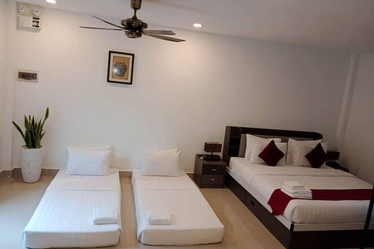 Habitación de hotel con dos camas individuales, una cama más grande, un ventilador de techo, una planta en maceta y un aplique de pared.