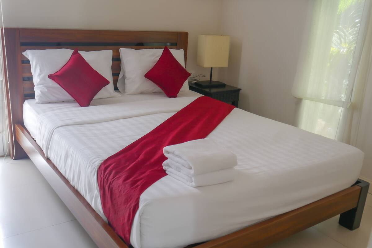 Una cama bien hecha con sábanas blancas y detalles rojos, un cabecero de madera y una mesita de noche con una lámpara.