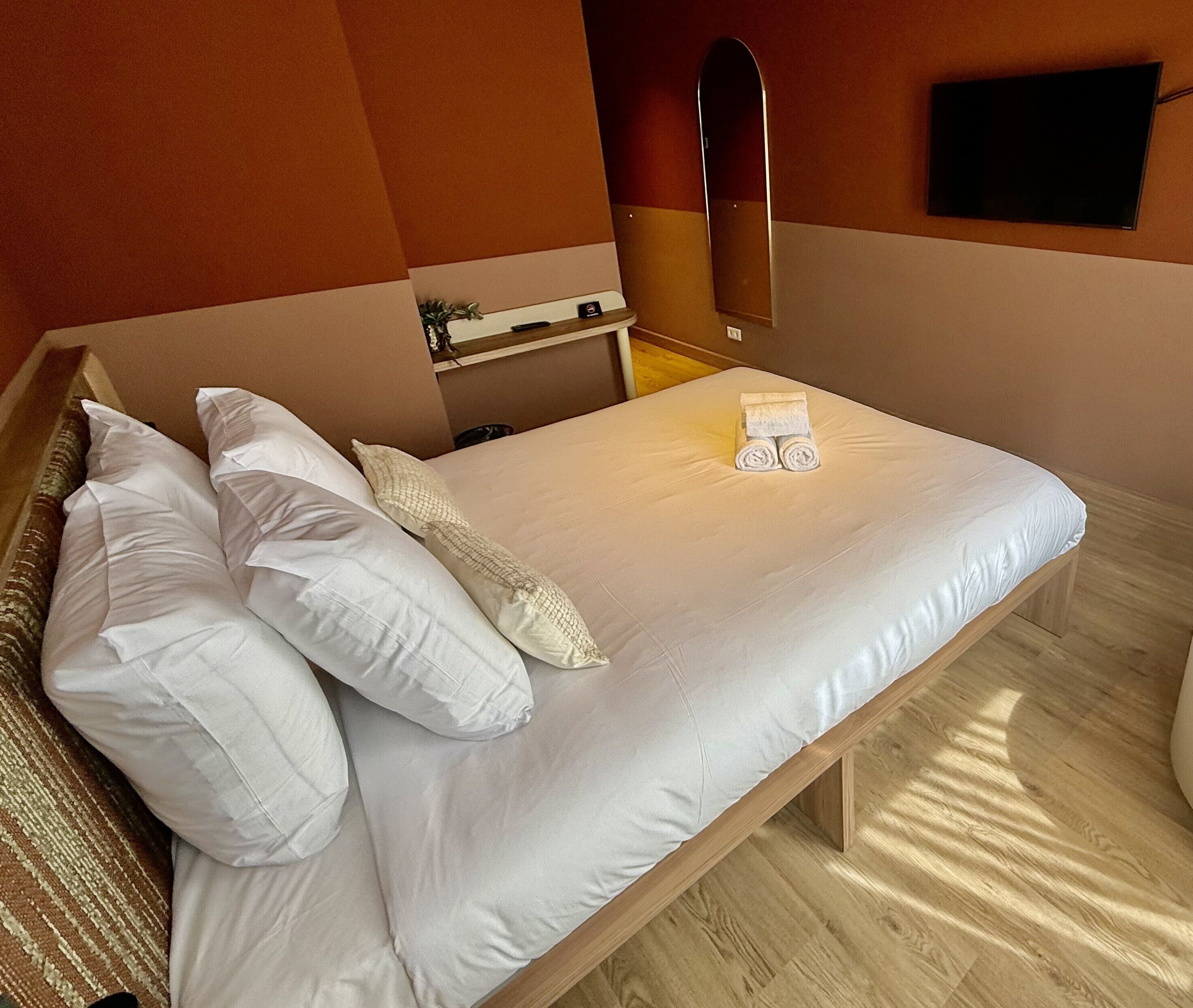 Una cama bien hecha con sábanas y almohadas blancas, un cabecero de madera, una mesita de noche con una planta y un televisor de pantalla plana montado en la pared.