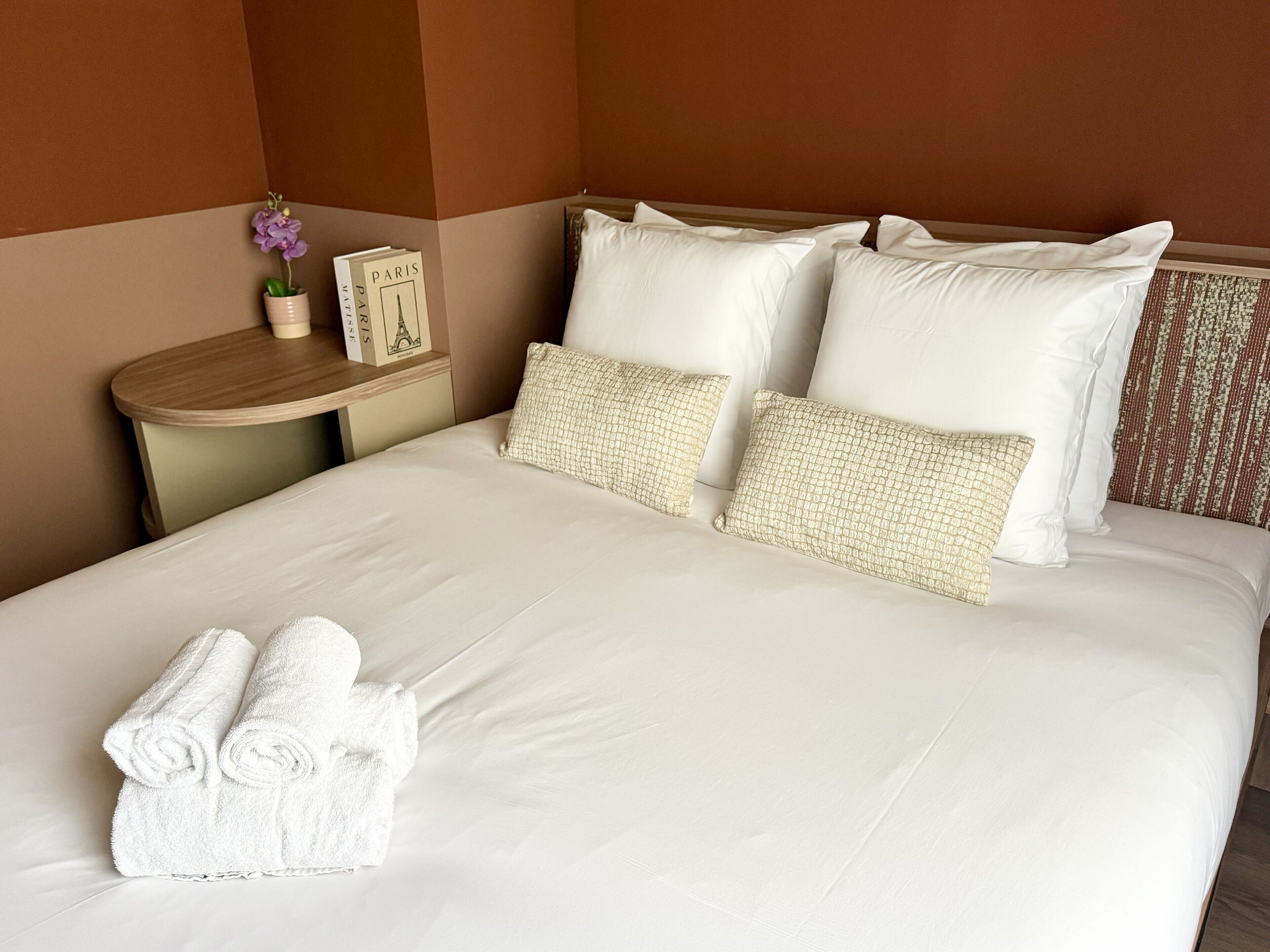 Una cama bien hecha con sábanas blancas y toallas enrolladas, una mesita de noche con un florero y un libro titulado 'PARIS', y un cabecero estampado.