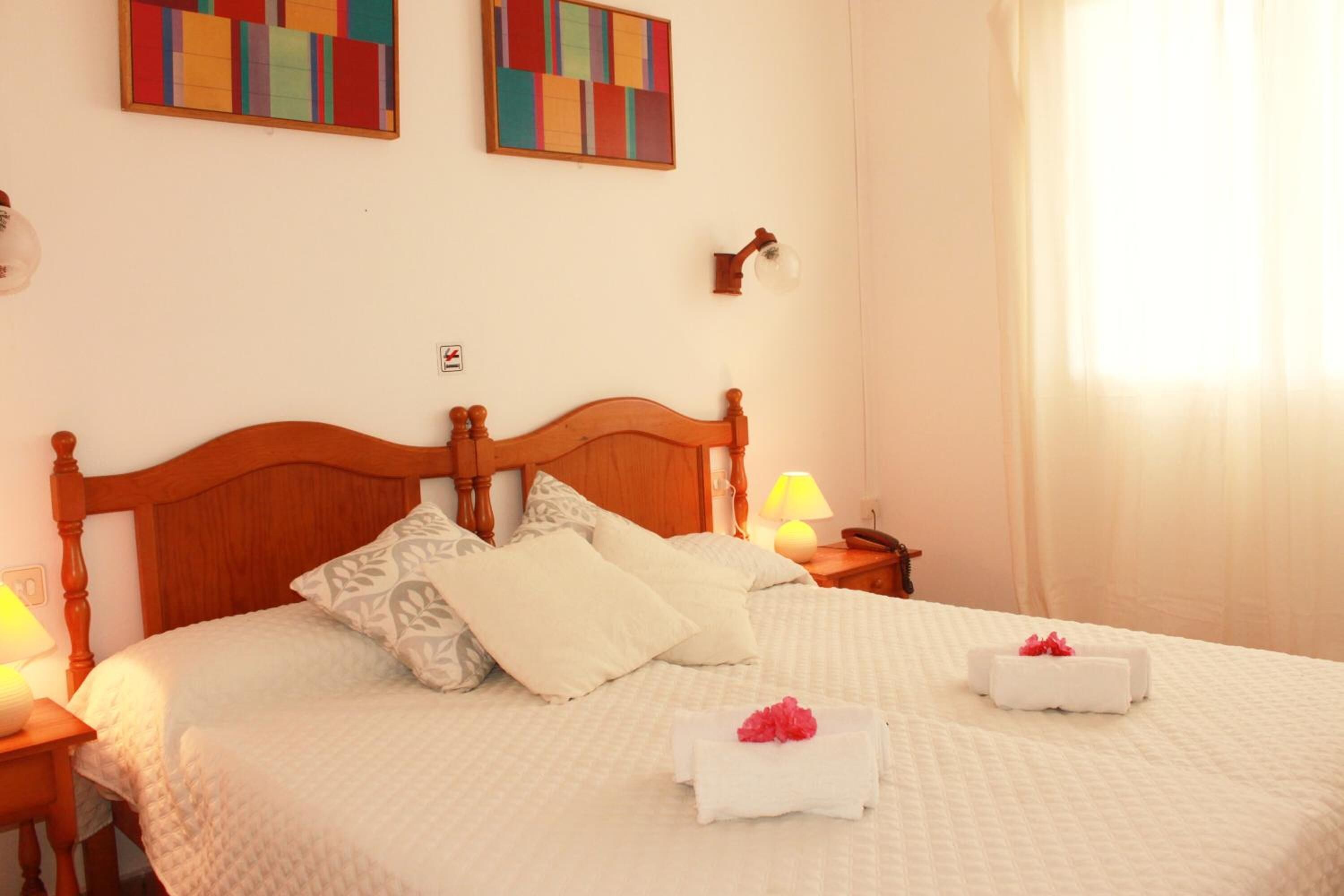 Un letto rifatto con cura, con testiera in legno, biancheria bianca e due asciugamani con fiori rosa.