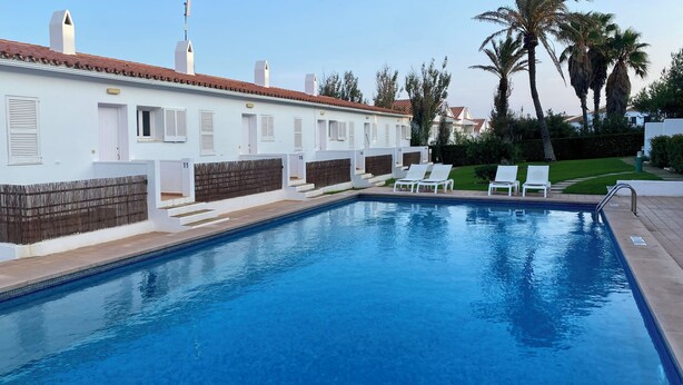 Una piscina con sillas de descanso, un edificio blanco con puertas numeradas y palmeras al fondo.