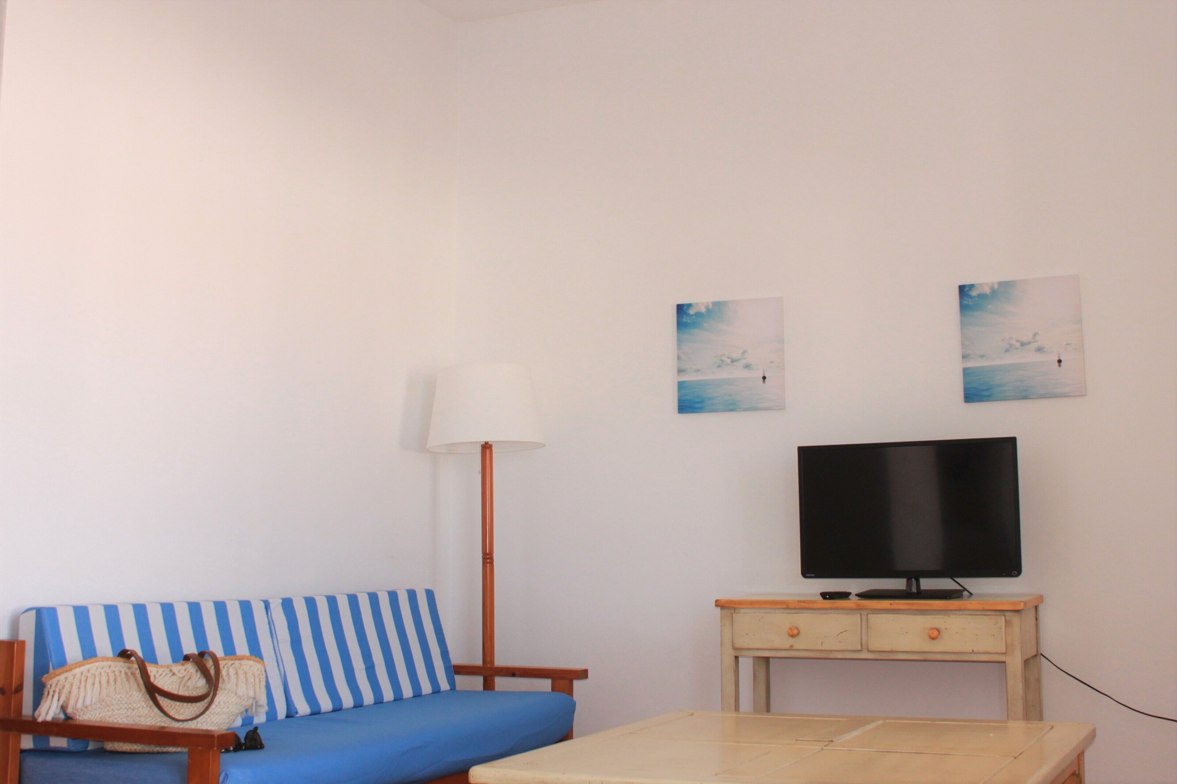 A room with a blue and white striped sofa, a television on a wooden stand, a lamp, and two wall-mounted pictures.
