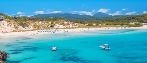 A beach with clear turquoise waters, boats, and a mountainous backdrop.