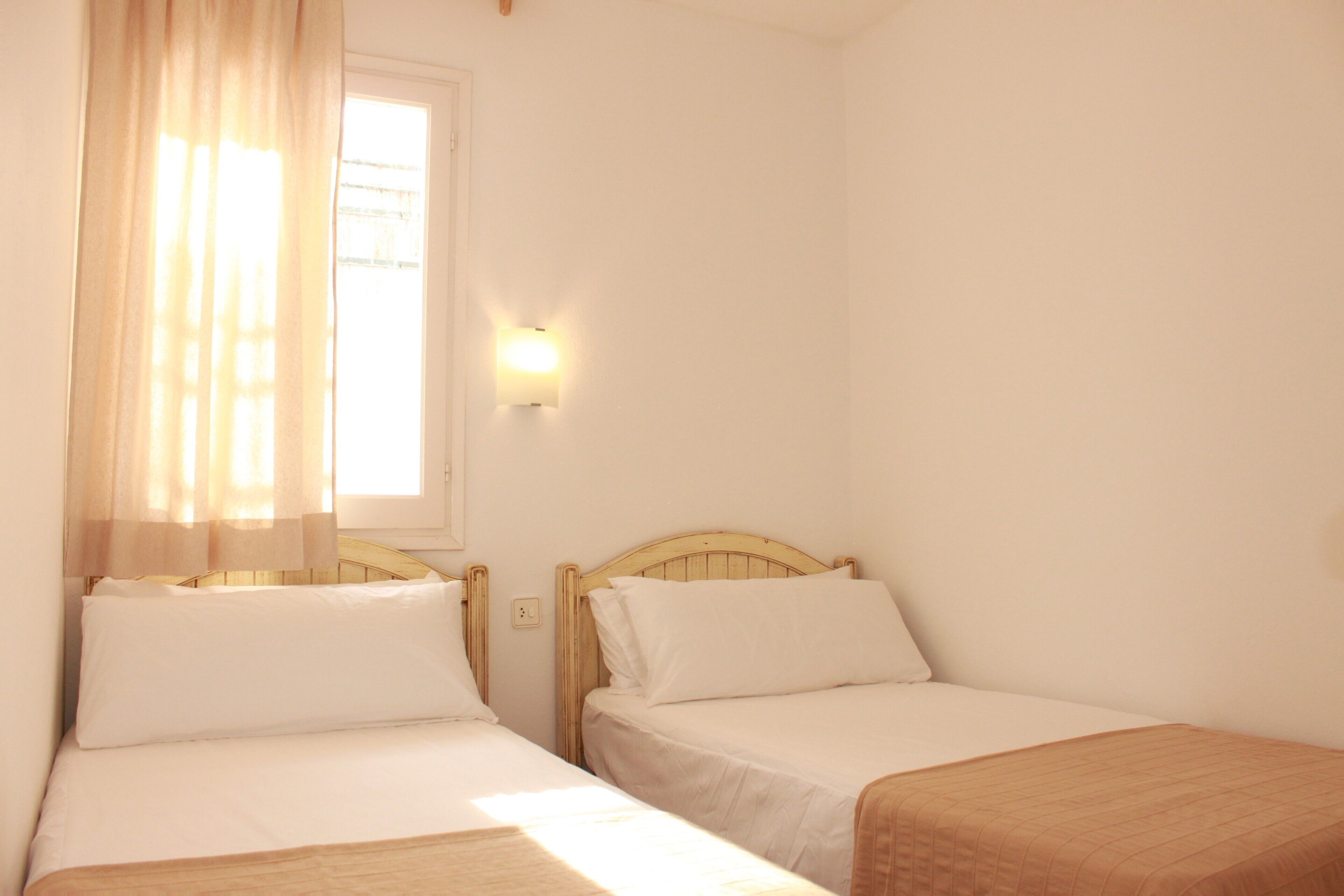 A room with two single beds, a window with sheer curtains, a wall-mounted light, and a plain white wall.