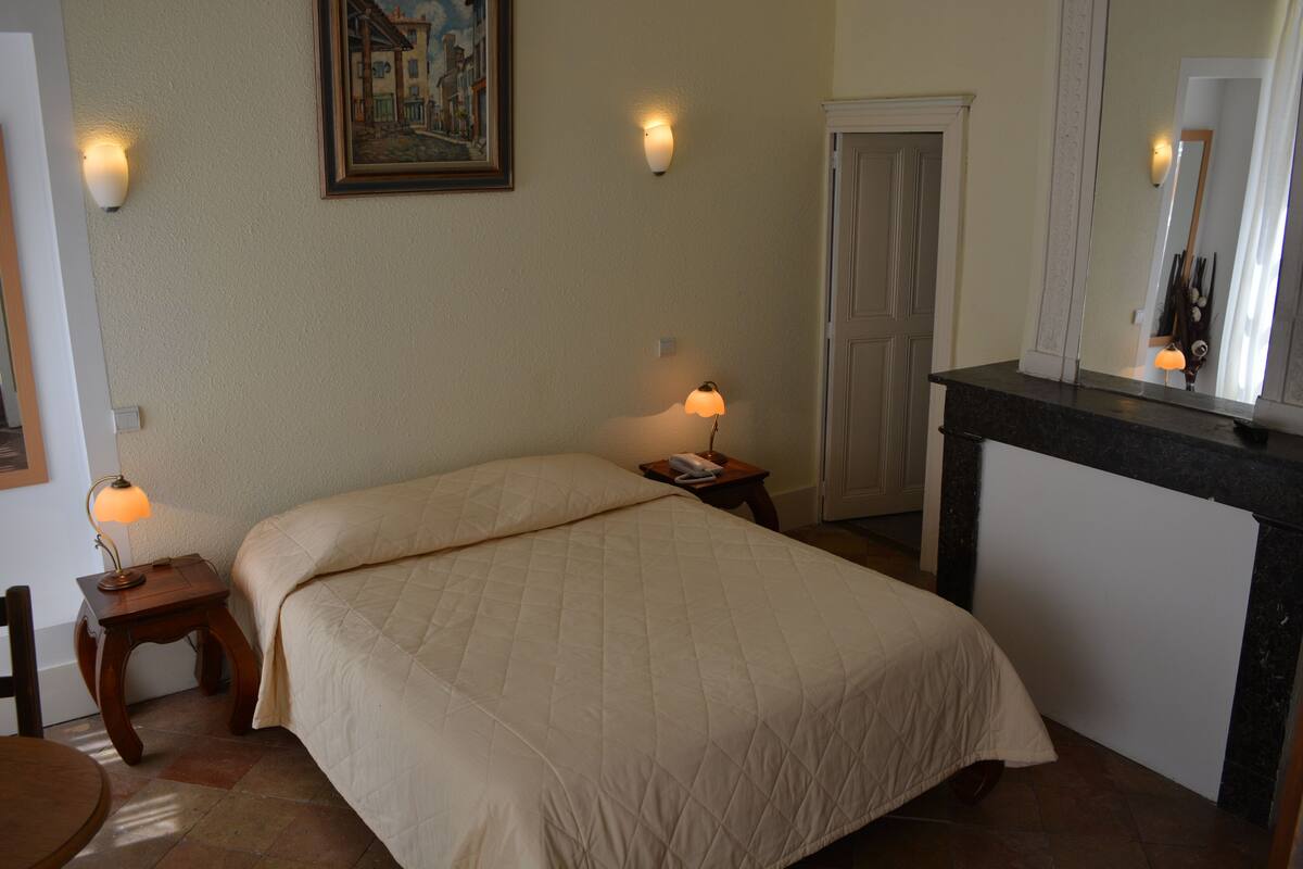 Ein ordentlich bezogenes Bett mit einer Steppdecke, zwei Nachttischlampen, ein kleiner Tisch mit Lampe, ein Gemälde an der Wand und ein Spiegel über einer dunkelbraunen Kommode.