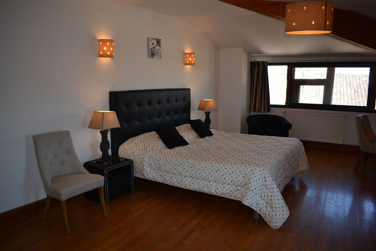 Ein Schlafzimmer mit einem großen Bett, zwei Nachttischlampen, einem Sessel und einem Vorhangfenster.