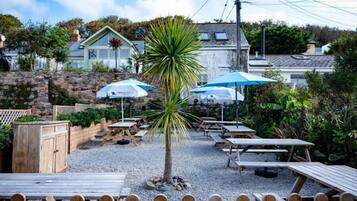 An outdoor seating area with wooden tables and blue umbrellas, surrounded by a stone wall and greenery.