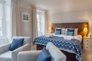 Una habitación de hotel con una cama grande, dos mesitas de noche con lámparas, una silla y un ventanal con cortinas.