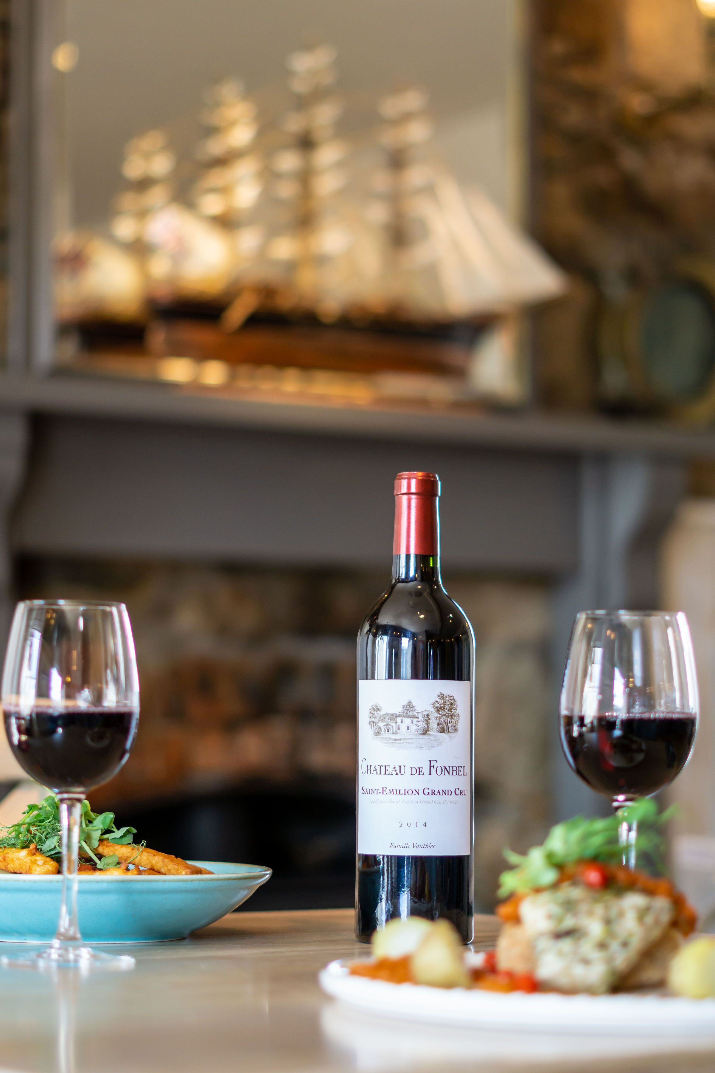A bottle of Chateau de Fonbel wine, two glasses of red wine, and a plate of food on a table.