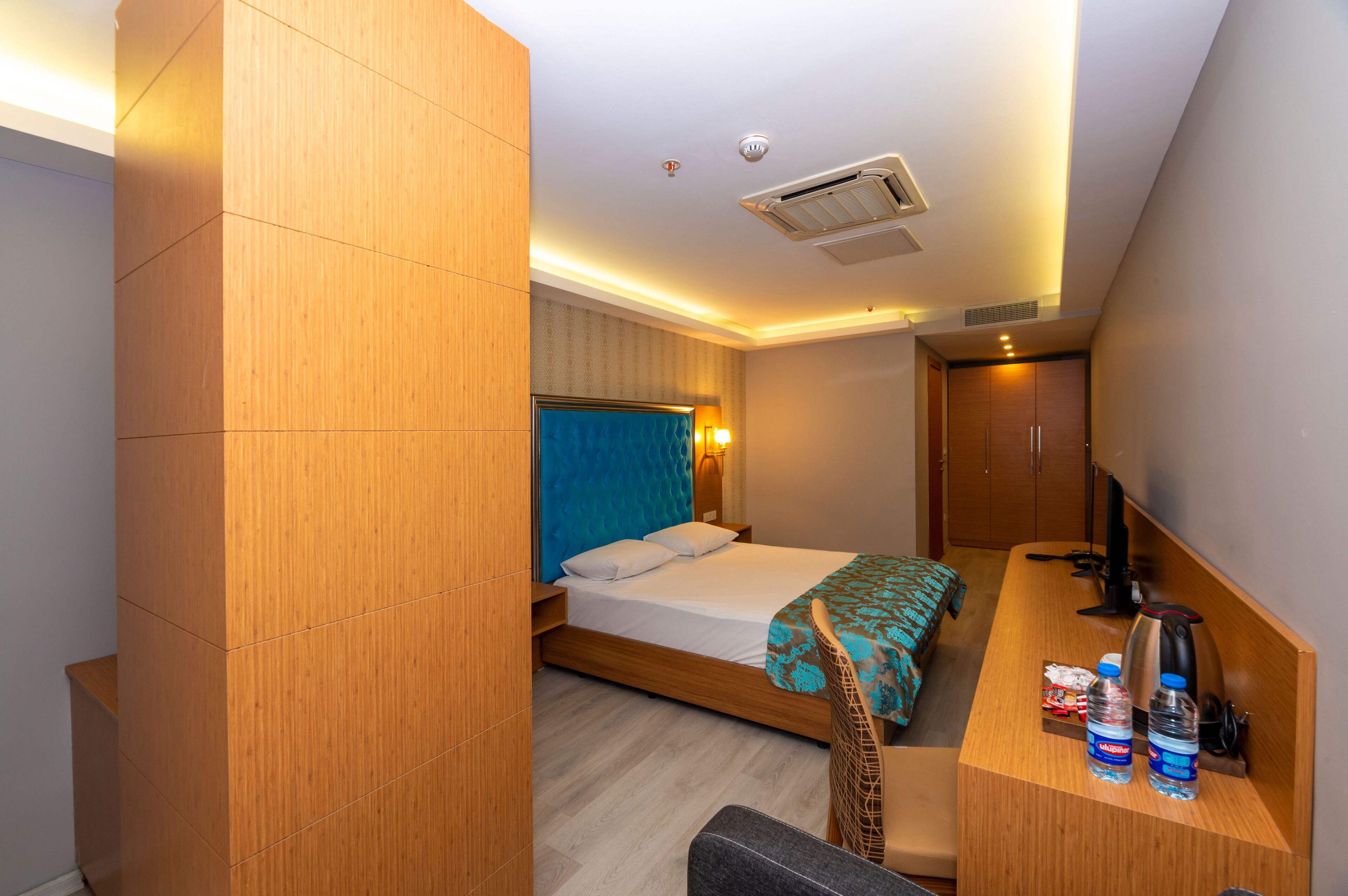 A modern hotel room with a wooden bed, a desk with a chair, a television, and a minibar.