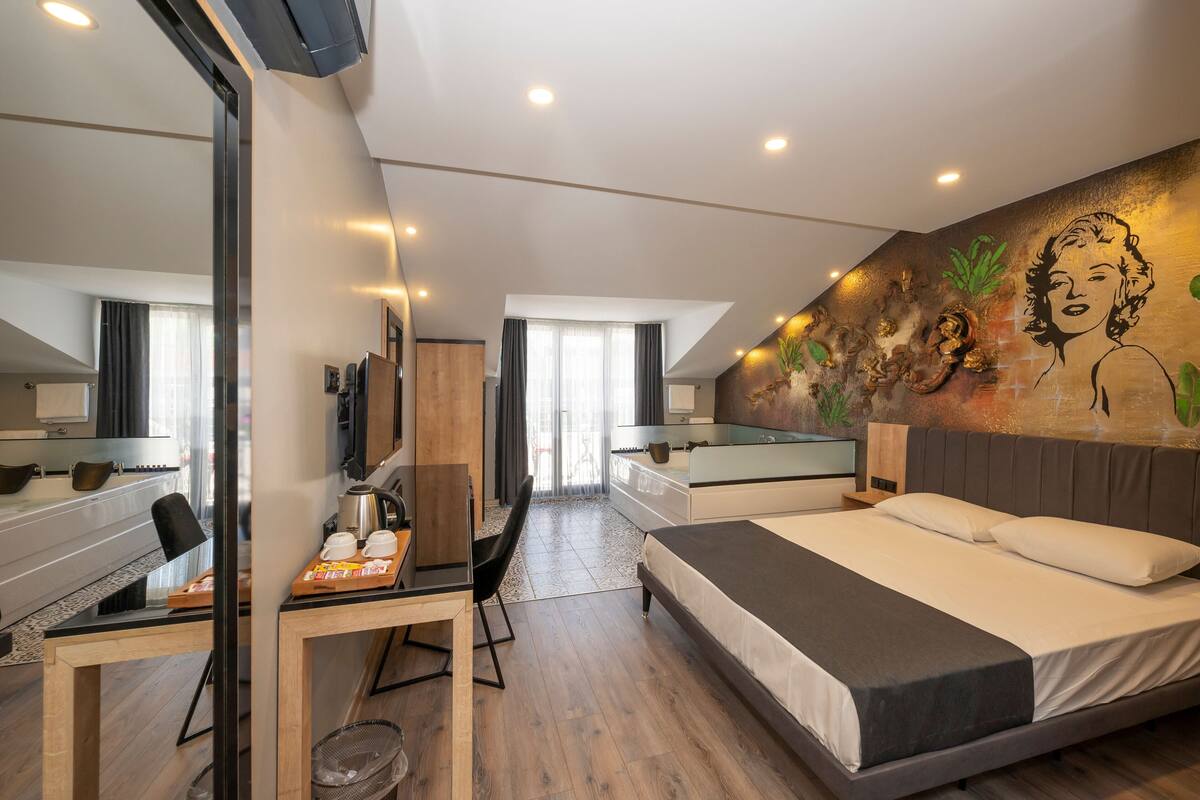 A modern hotel room with a large bed, a desk with a chair, a small table, and a large mirror.