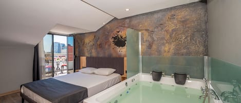 A modern hotel room with a large bed, a separate sleeping area, a bathtub, and a city view through large windows.