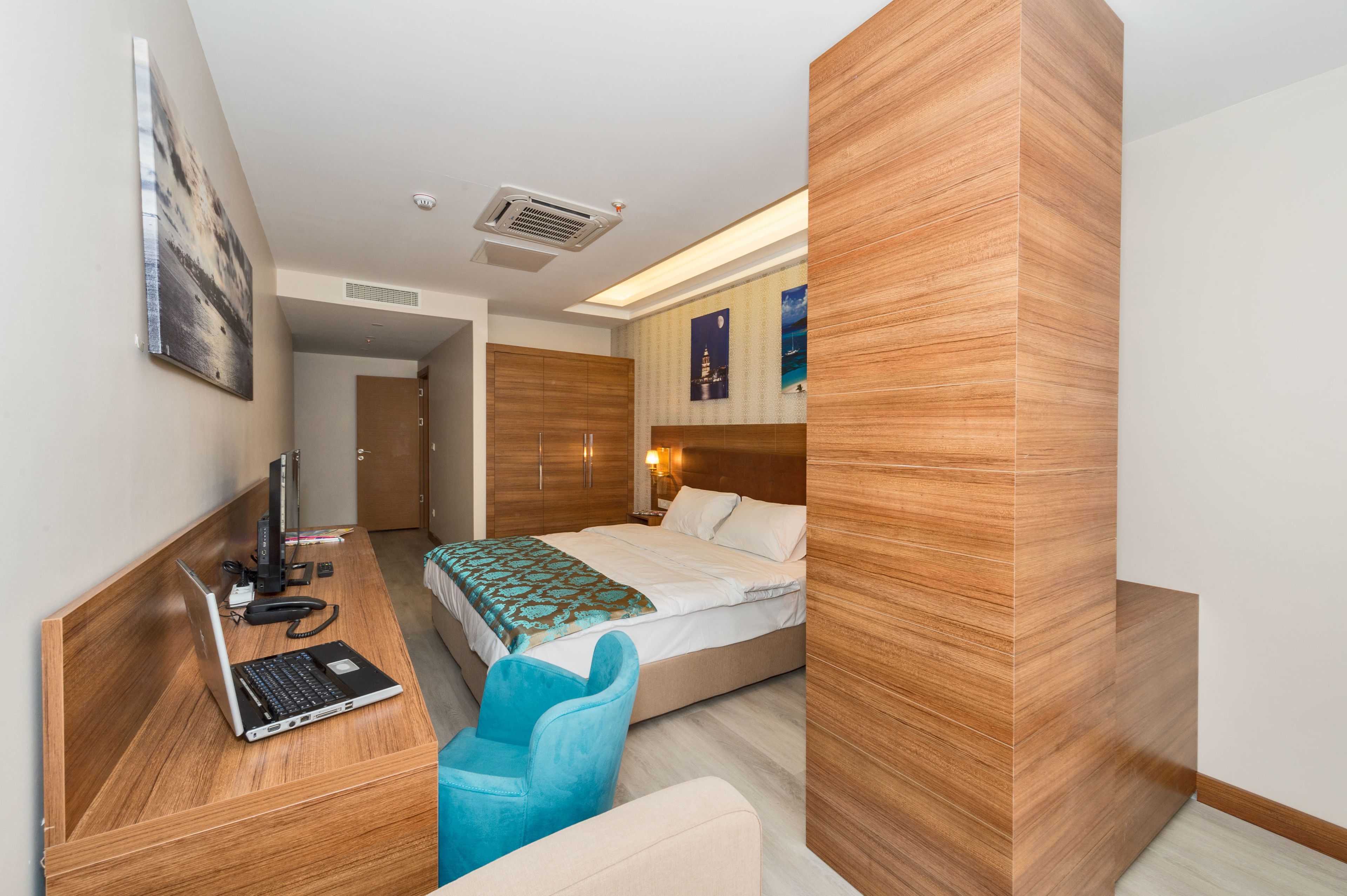 A hotel room with a wooden desk, a laptop, a blue chair, a bed, and a nightstand.