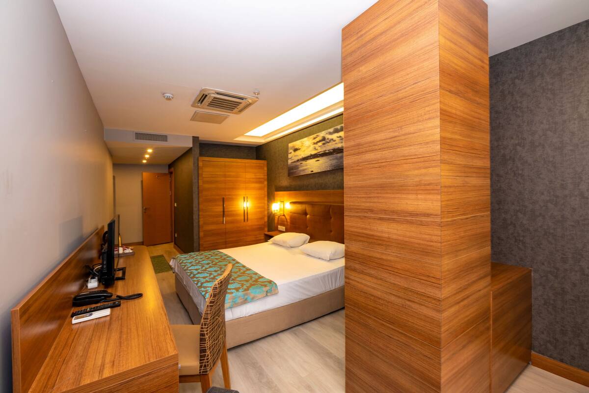 A modern hotel room with a wooden desk, a bed with a blue and white patterned throw, a headboard with built-in lighting, and a large artwork on the wall.