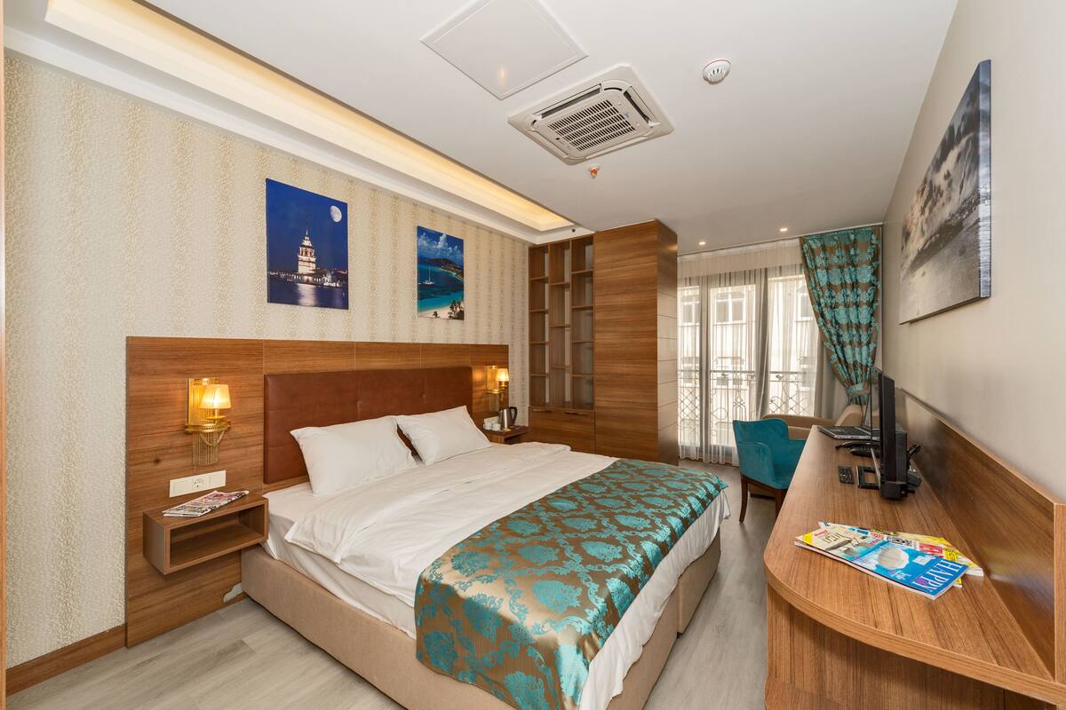 A hotel room with a large bed, a wooden headboard, a nightstand with a lamp, a desk with a computer, and a window with curtains.
