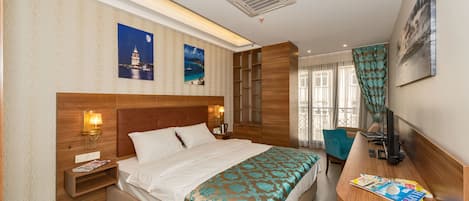 A hotel room with a large bed, a wooden headboard, a nightstand with a lamp, a desk with a computer, and a window with curtains.