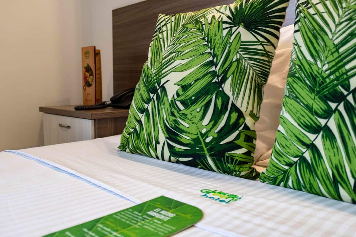 Une chambre d’hôtel avec un lit agrémenté d’oreillers à motifs de feuilles vertes, un lit parfaitement fait avec un drap blanc, et une brochure verte de l’hôtel posée sur le lit.