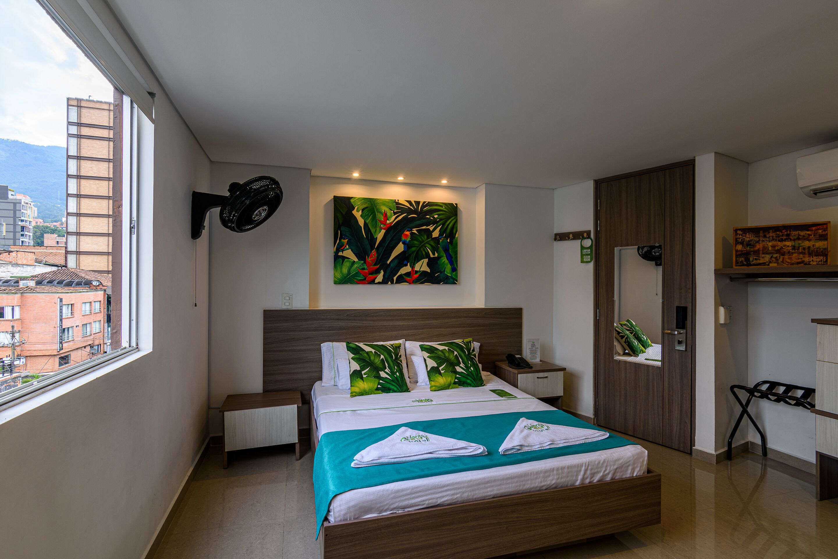 Quarto de hotel moderno com cama, mesa de cabeceira, ventoinha na parede, pintura e vista da cidade pela janela.