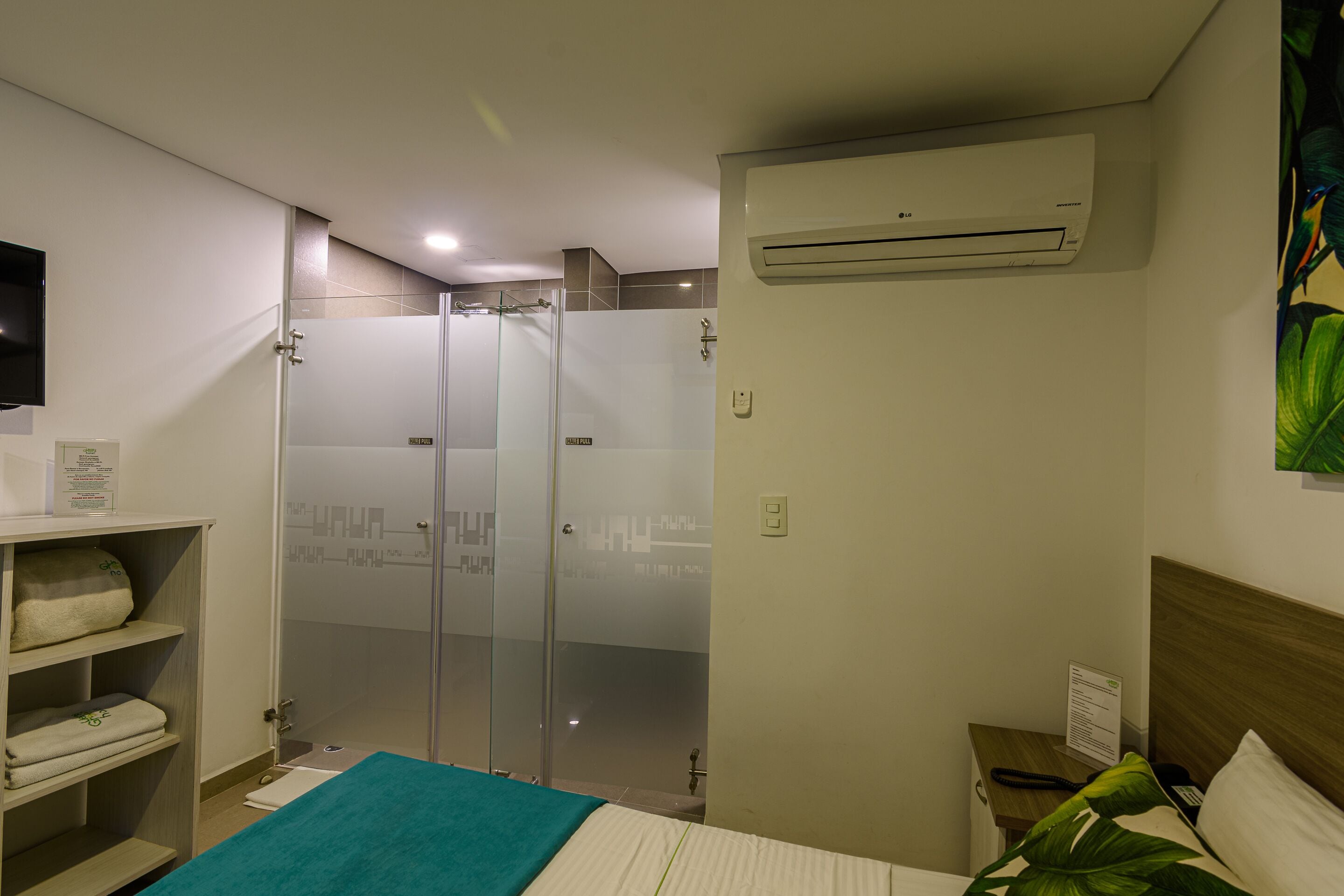 A hotel room with a bed, a shower area, a shelf with towels, an air conditioning unit, and a wall-mounted TV.