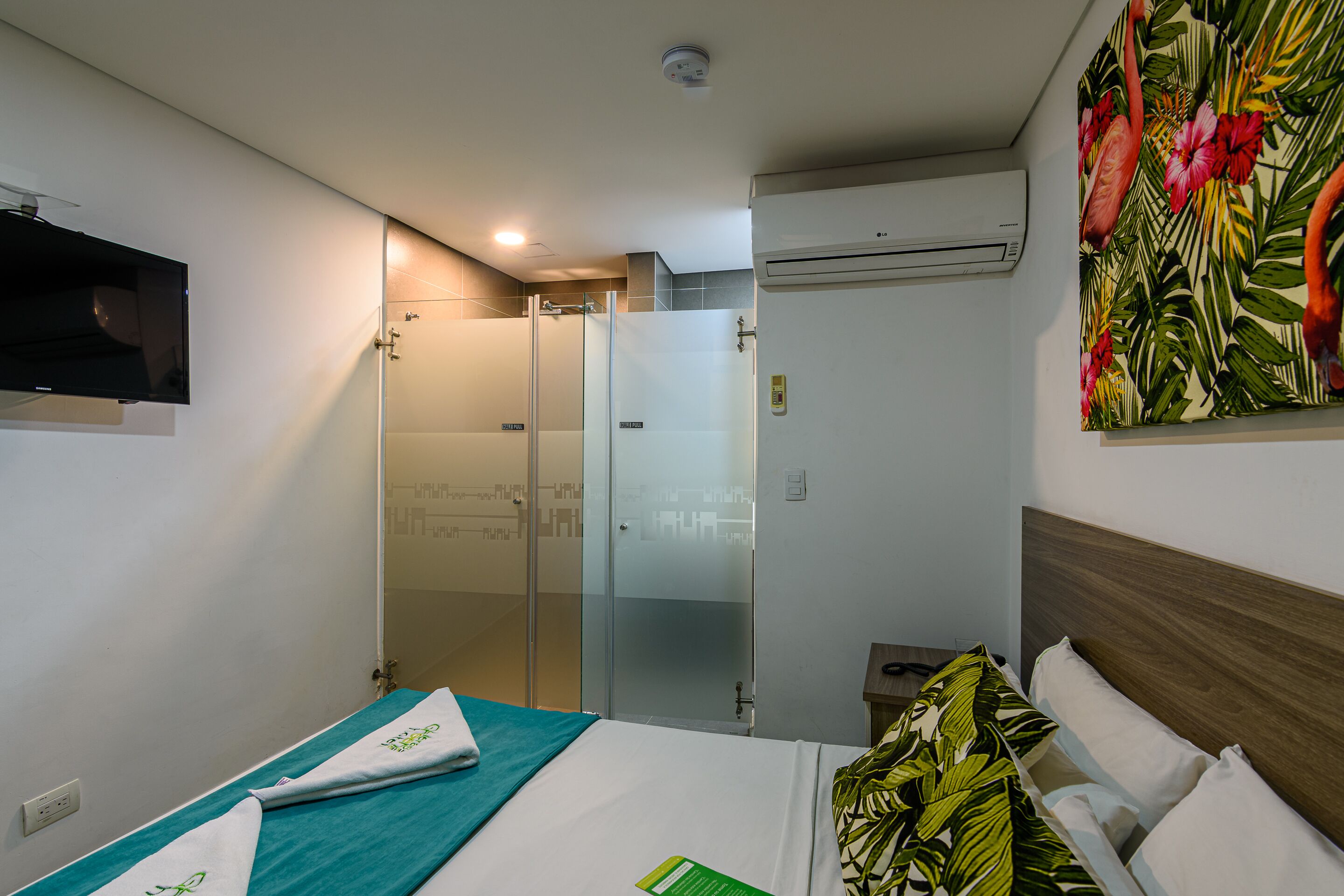 A hotel room with a bed, a TV mounted on the wall, a shower area, and a wall-mounted air conditioner.