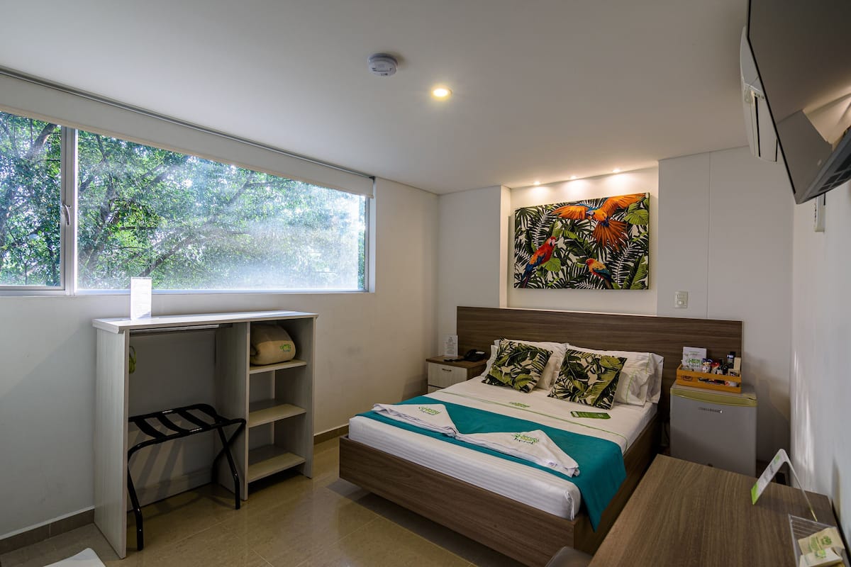 Une chambre d’hôtel avec un lit, un bureau, une chaise, un petit placard, une fenêtre et un tableau au mur.