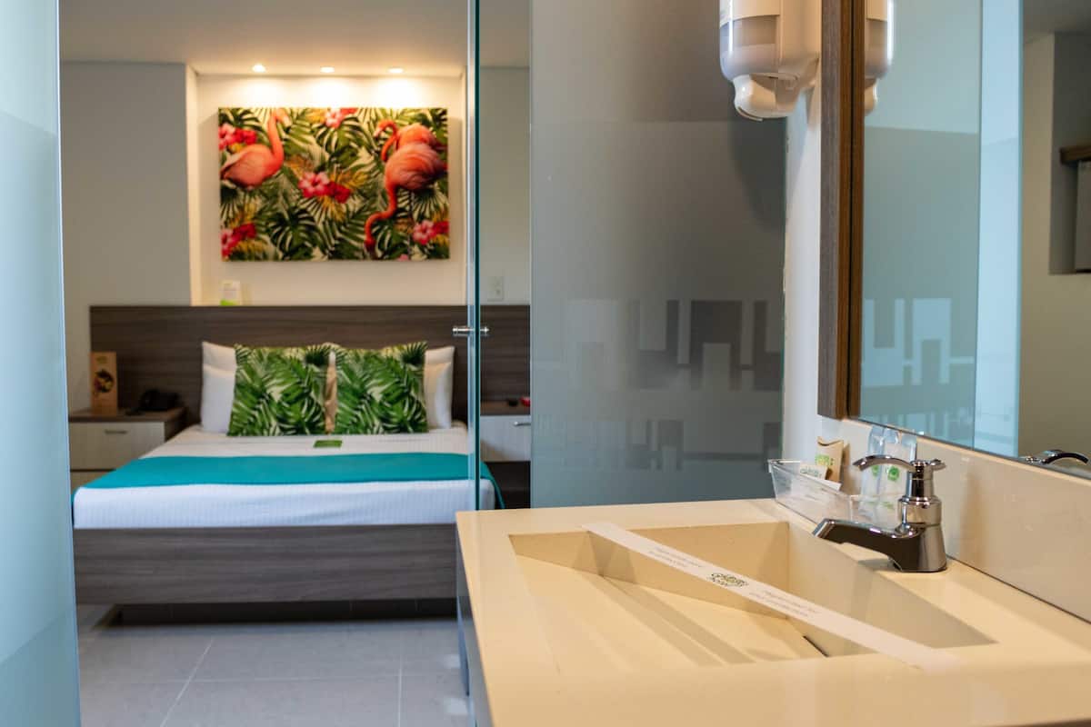 Une chambre d’hôtel avec un lit, une salle de bain dotée d’un lavabo et d’un miroir, et un tableau aux motifs tropicaux accroché au mur.