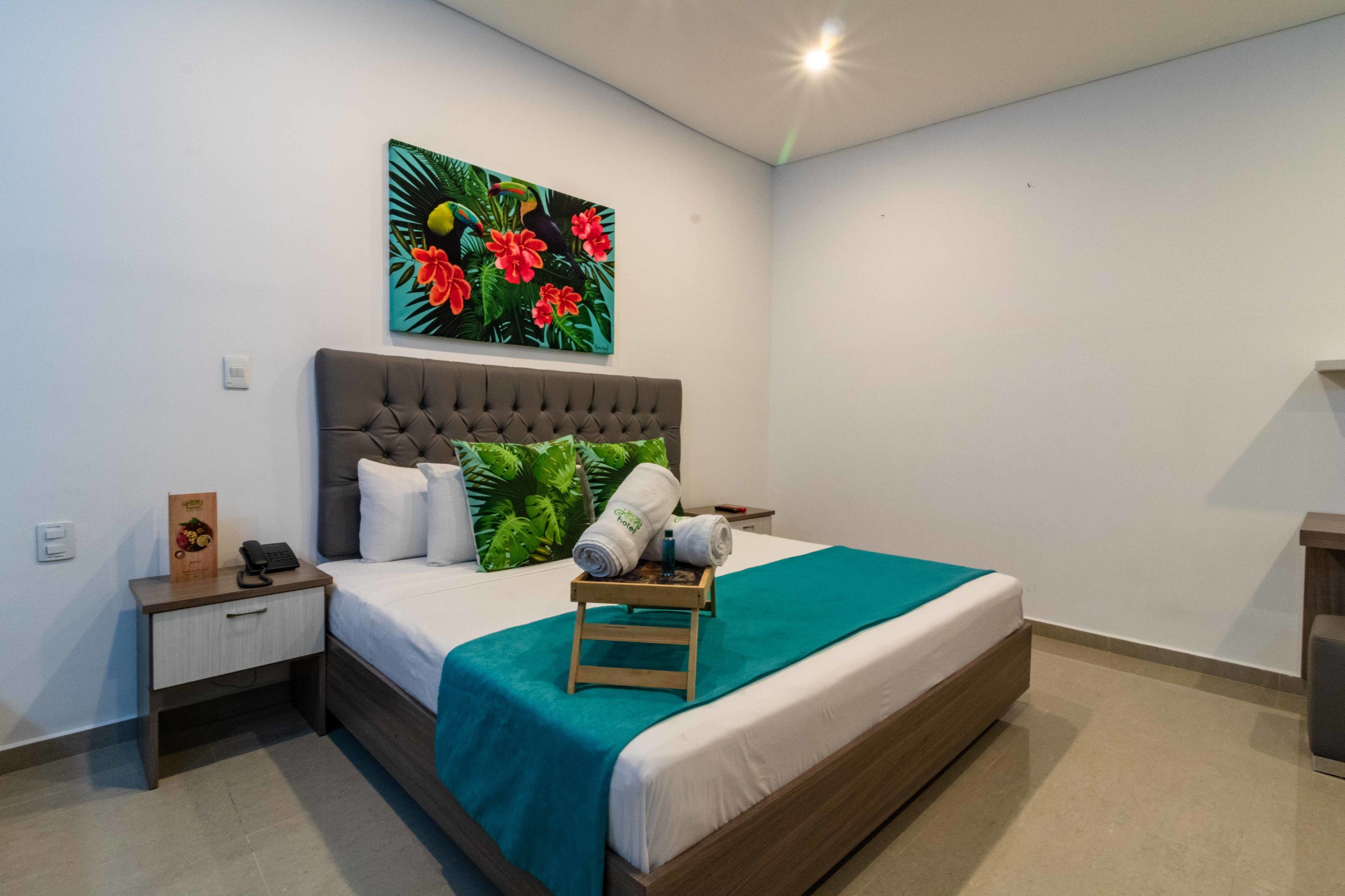 A neatly arranged hotel room with a bed, a nightstand, a wall-mounted painting, and a small wooden stool.