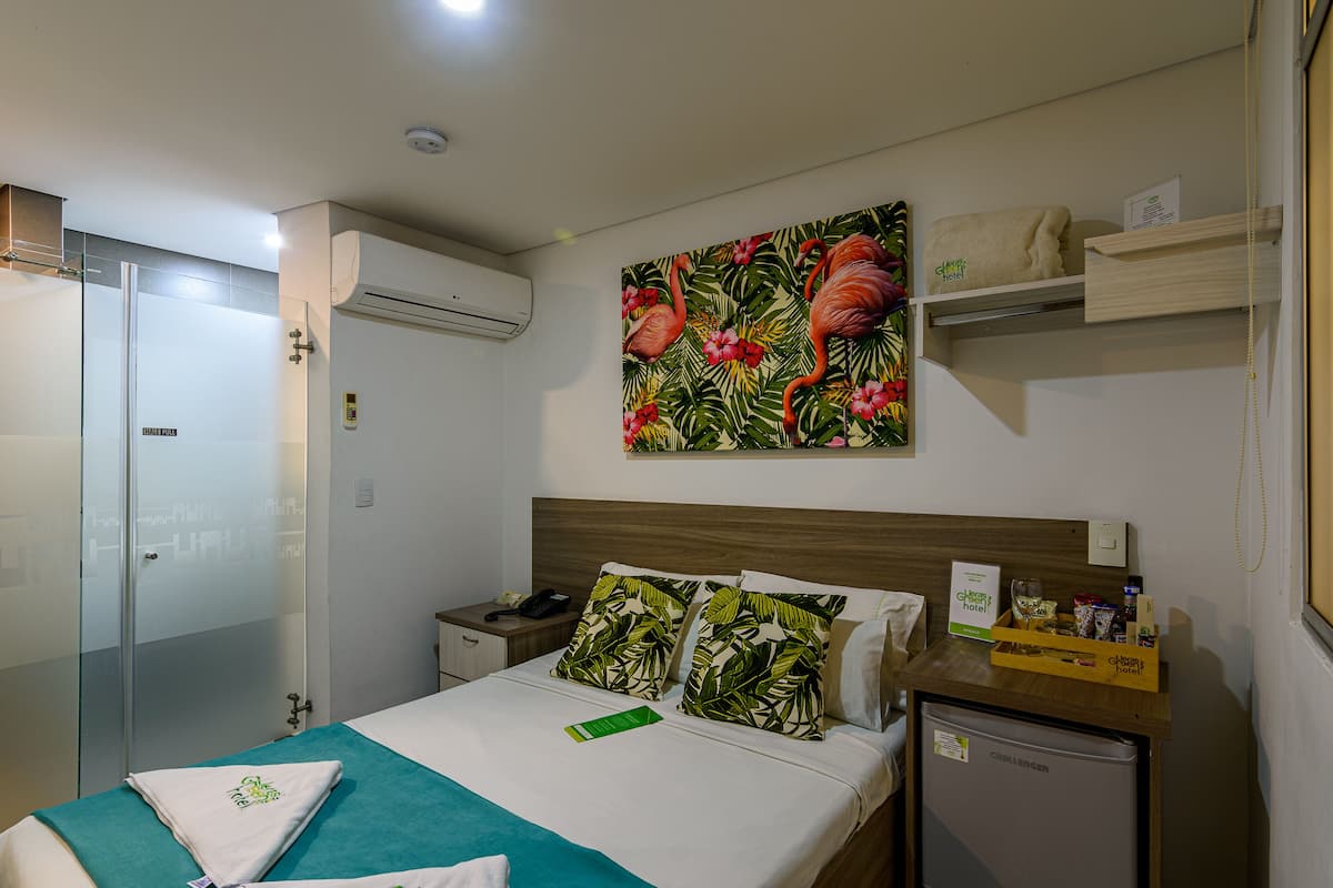 Une chambre d’hôtel comprenant un lit, une table de chevet, un mini-réfrigérateur, une serviette, un tableau et un espace douche.