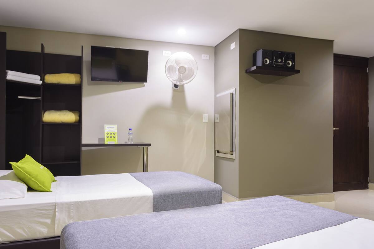 A hotel room with two beds, a TV, a fan, a small desk with a phone, and a door.